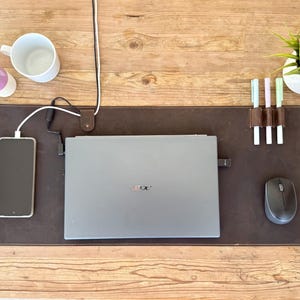 May include: A brown leather desk mat with a laptop, smartphone, and wireless mouse. The mat has a pen holder with three pens and a small plant. A white mug and pencil holder are also on the wooden desk.
