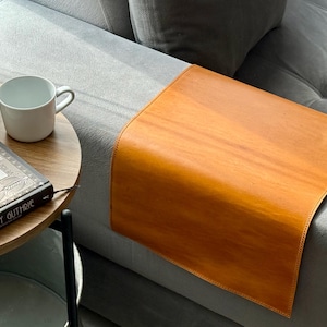 May include: A light brown leather sofa arm tray sits on a grey sofa. A white mug and a book titled "Woody Guthrie" are on a small, round wooden side table. The tray has rounded corners and a stitched edge.