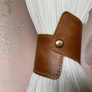 May include: Brown leather curtain tieback with a metal button. The tieback is wrapped around a white sheer curtain.