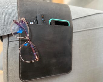 May include: A dark gray leather couch caddy draped over a light gray fabric sofa armrest. The organizer features a deep pocket containing two black remote controls and a smartphone with a teal case, with a pair of glasses resting on the front.