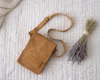 May include: A tan leather crossbody bag with a flap closure and adjustable strap lies on a white quilted surface. A bundle of dried lavender and a cream-colored knitted blanket are also present.