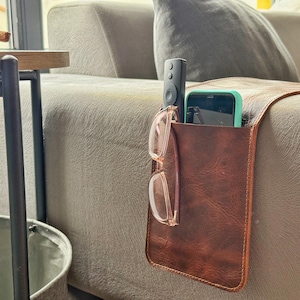 May include: A brown leather couch arm organizer with a pocket holding a remote, a smartphone, and eyeglasses. The organizer is designed to hang over the arm of a sofa, providing convenient storage for living room essentials.