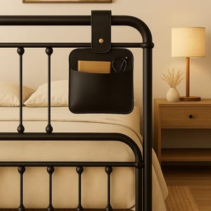 Può includere: Organizer da comodino in pelle nera appeso a una struttura letto in metallo nero. L'organizer ha una tasca con un oggetto color oro e un paio di occhiali. Un piccolo bottone dorato fissa il cinturino. Il letto ha una trapunta beige.
