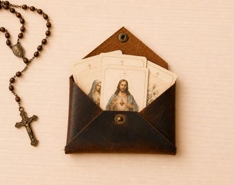 May include: A dark brown leather envelope-style pouch with a metal snap closure containing several cream-colored prayer cards featuring religious illustrations of Jesus and Mary, accompanied by a brown wooden rosary with a metal crucifix.