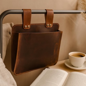 May include: A brown leather bedside organizer hanging from a metal bed frame. The organizer has two straps with metal snaps and a large pocket. A cup of coffee and an open book are on the nightstand.