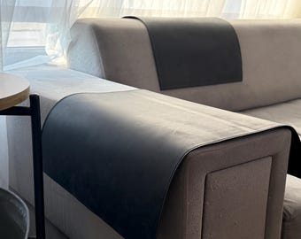 May include: A rectangular black protective cover draped over the armrest of a light gray sofa. The smooth, dark material extends across the top and down the side of the armrest, providing a contrast against the light-colored upholstery.