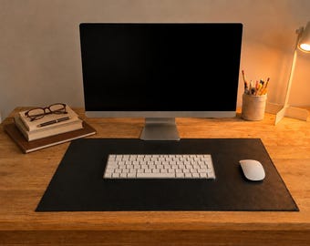May include: A computer monitor with a black screen, a white wireless keyboard, and a white computer mouse placed on a black rectangular desk mat. A stack of books with glasses and a pen, a pencil holder, and a white desk lamp are also present.