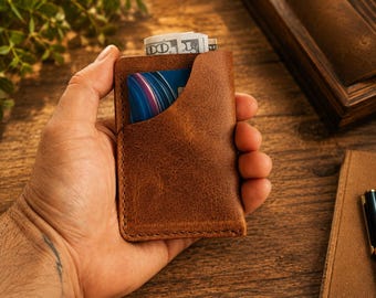 May include: A tan brown leather card holder wallet featuring visible stitching along the edges, a curved top pocket containing a blue and purple patterned credit card, and a top slot holding folded United States one hundred dollar bills.