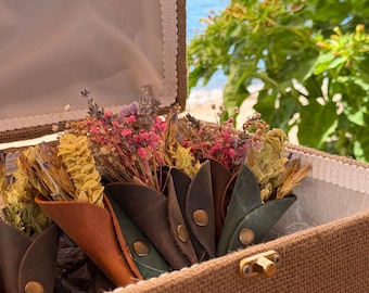 May include: A burlap-lined box containing several small, cone-shaped leather holders in shades of brown, tan, and green, each secured with a brass snap button. Inside each leather cone is a bouquet of dried flowers in pink, yellow, and purple.