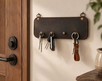 May include: A rectangular dark brown leather wall-mounted key holder featuring four brass-colored metal hooks. The leather panel is secured with visible stitching along the edges and includes two small metal rings at the top for hanging purposes.