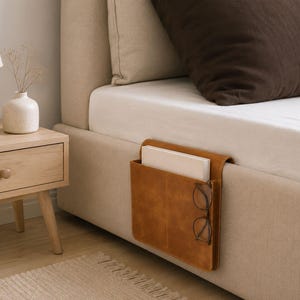 May include: A brown leather bedside caddy with a pocket holding books and eyeglasses. The caddy is attached to the side of a bed with a beige headboard. A wooden nightstand with a lamp and vase is in the background.