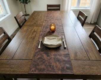 May include: A dark brown wooden dining table set for a meal. A brown leather table runner is centered on the table, with a plate, napkin, fork, knife, and candle. The table is surrounded by wooden chairs.