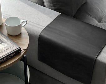 May include: A black leather sofa arm tray draped over the arm of a gray upholstered sofa. The tray provides a flat surface for items. Nearby is a round wooden side table holding a white ceramic mug and a book titled Woody Guthrie.