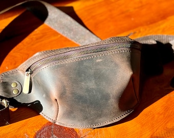 May include: A brown leather fanny pack with a zipper closure and adjustable strap. The bag has a distressed finish and brass-colored hardware. The fanny pack is designed to be worn around the waist or across the body.