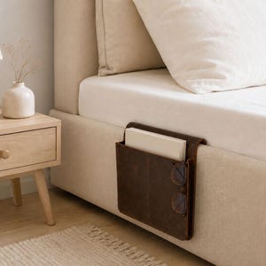 Puede incluir: Un organizador de cama de cuero marrón con dos compartimentos, que contiene un libro y gafas. El organizador está sujeto al lateral de una cama beige. Una mesita de noche de madera con una lámpara y un jarrón está en el fondo.