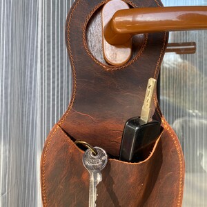 May include: A brown leather key holder with a pocket for keys and a small pocket for a phone or other small items. The key holder is hanging on a door handle.