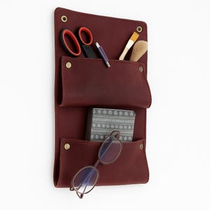 Könnte beinhalten: Ein weinroter Leder-Wandorganizer mit drei Taschen, der eine Schere, einen Stift, Pinsel, eine kleine gemusterte Schachtel und eine Brille enthält. Der Organizer hat Messingnieten und ist an einer weißen Wand befestigt.