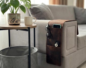 May include: A brown leather couch armrest organizer with multiple pockets, holding a phone, remote, and sunglasses. A small side table with a plant and mug is next to the couch. A gray basket sits below the table.
