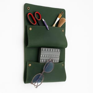 Könnte beinhalten: Ein dunkelgrüner Wandorganizer aus Leder mit drei Taschen. Die obere Tasche enthält eine rotgriffige Schere, einen Stift und einen Pinsel. Die mittlere Tasche enthält eine gemusterte Box. Die untere Tasche enthält eine Brille. Nieten sichern den Organizer.