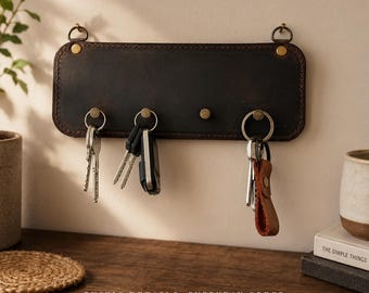 May include: A rectangular dark brown leather wall-mounted key holder featuring four brass-toned metal hooks, two top mounting rings, and visible stitching along the edges. Three of the hooks hold various metal keys and a brown leather keychain.