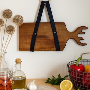 May include: A wooden cutting board with an antler design, hanging on a white wall. It has a dark blue leather strap and a wooden ring. The board is approximately 46 cm long. Kitchenware.