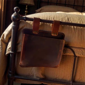 May include: A dark brown leather bed organizer with two brown leather straps attached to a metal bed frame. The organizer has a pocket for storage. The bed frame is dark brown and the bedding is tan.