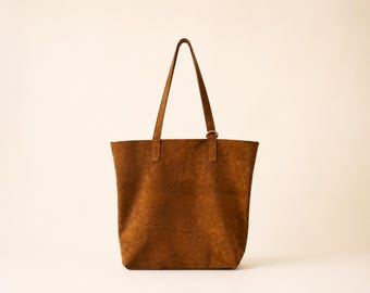 May include: A brown suede tote bag with a rectangular body and two long shoulder straps. The bag features a simple, unadorned design with a single metal ring attachment on one of the straps near the top edge, providing a clean and functional look.