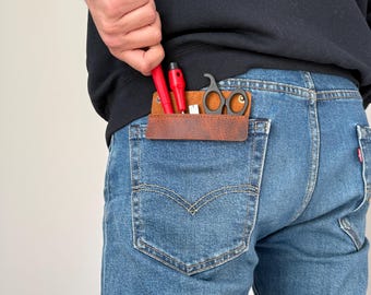 May include: A brown leather pocket organizer with tools, including red screwdrivers and small black scissors, is visible in the back pocket of blue jeans. The person is wearing a black sweatshirt.
