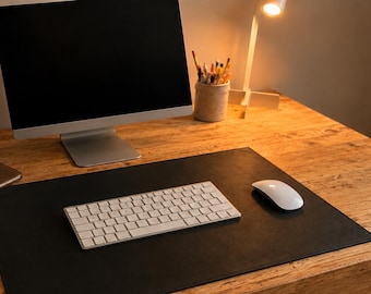 May include: A black rectangular desk mat made of a smooth material, positioned on a wooden surface. The mat serves as a base for a white wireless keyboard and a white computer mouse, with a computer monitor and a pencil holder nearby.