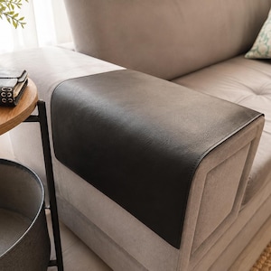 May include: A rectangular black faux leather armrest cover draped over the arm of a light gray upholstered sofa. The cover has a smooth, matte texture and fits snugly over the top and sides of the sofa arm, providing a protective layer.
