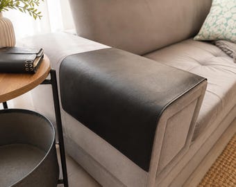 May include: A rectangular black faux leather armrest cover draped over the arm of a light gray upholstered sofa. The cover features a smooth, matte texture and is positioned to protect the top surface of the sofa arm from wear and tear.
