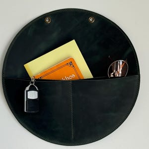 Könnte beinhalten: Ein dunkelgrüner, kreisförmiger Wandorganizer aus Leder mit zwei Taschen. Er enthält einen gelben Notizblock, ein kleines oranges Buch, eine Sonnenbrille und einen Schlüsselbund. Der Organizer wird mit zwei Messingnieten an der Wand befestigt.