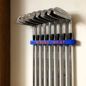 May include: A set of Mizuno MP-67 golf clubs, with shiny silver heads and black grips, held in a blue holder. The holder displays the text "MIZUNO MP-67" in orange. The clubs are arranged vertically.