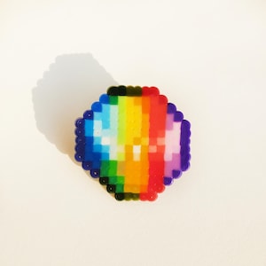 Peut inclure: Une épingle en forme de cœur arc-en-ciel pixélisée. L'épingle présente un spectre de couleurs, notamment le bleu, le vert, le jaune, l'orange et le rouge. Les bords sont soulignés en bleu foncé et en noir.