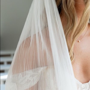 May include: A sheer white wedding veil with delicate floral lace detailing. The veil is draped over the shoulder, revealing a glimpse of the bride's dress and blonde hair. A gold necklace with a pearl pendant is visible.