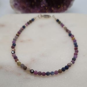 May include: A delicate bracelet with small, faceted beads in shades of purple, blue, brown, and silver. The beads are strung together with a silver clasp. The bracelet is displayed on a white surface.