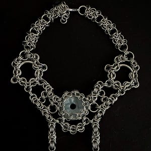 May include: A silver chainmail necklace with a central circular design and two dangling spikes. The necklace features intricate woven chain links and a clasp closure. The central design has a grey stone. The necklace is displayed against a black background.