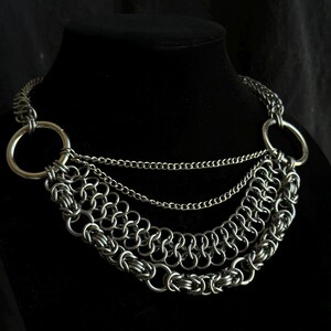 May include: A silver chain necklace with three layers of chain and two large silver rings. The necklace is on a black mannequin.