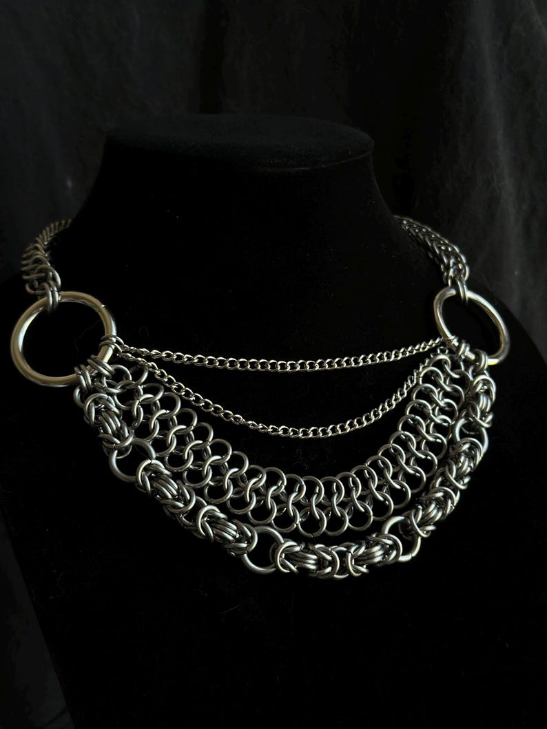 May include: A silver chain necklace with three layers of chain and two large silver rings. The necklace is on a black mannequin.