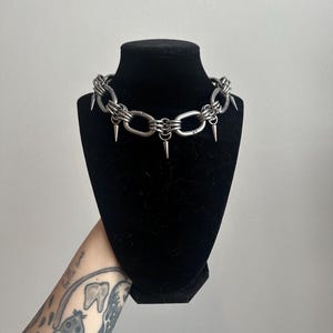 May include: A silver-colored choker necklace with a chain link design and multiple cone-shaped spikes. The necklace is displayed on a black mannequin head against a white background.