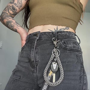 Może przedstawiać: Srebrny łańcuszek do kluczy przymocowany do czarnych dżinsów. Posiada karabińczyk, łańcuch i małą czarną metkę z białym tekstem. Do łańcuszka przymocowane są dwa klucze.