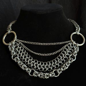 May include: A silver chain necklace with three layers of chain links and two large silver rings at the ends.