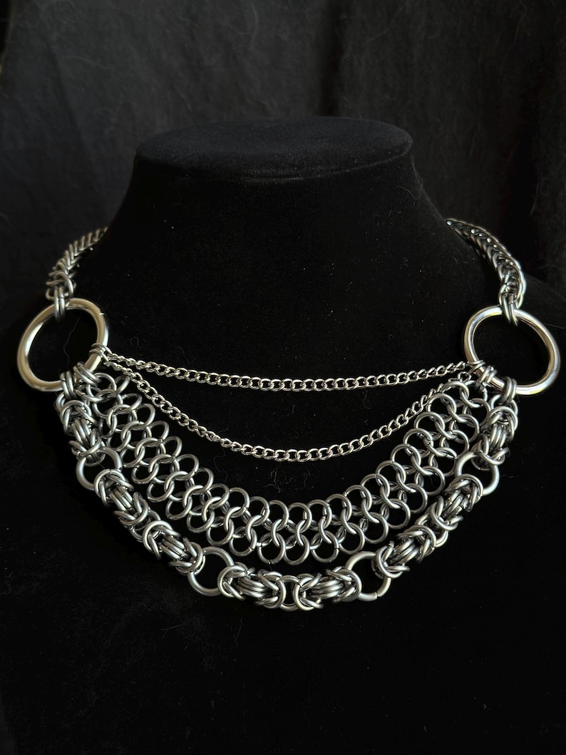 May include: A silver chain necklace with three layers of chain links and two large silver rings at the ends.