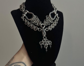 May include: A silver-colored chainmail necklace displayed on a black mannequin. The necklace features a series of interconnected rings forming a collar, with a central pendant of a three-pronged design. The necklace is intricate and detailed.