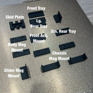 May include: A collection of black 3D-printed components, including a skid plate, trays, and various mounts. Text labels identify each piece, such as "Front Tray" and "Body Mag Mount." The parts are arranged on a textured, metallic surface.