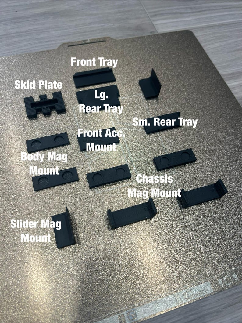 May include: A collection of black 3D-printed components, including a skid plate, trays, and various mounts. Text labels identify each piece, such as "Front Tray" and "Body Mag Mount." The parts are arranged on a textured, metallic surface.
