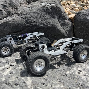 May include: Two miniature off-road vehicles with silver frames and black tires are positioned on a rocky surface. The vehicles have detailed suspension systems and silver wheels. One vehicle has a purple component. The scene is set outdoors with rocks and greenery.
