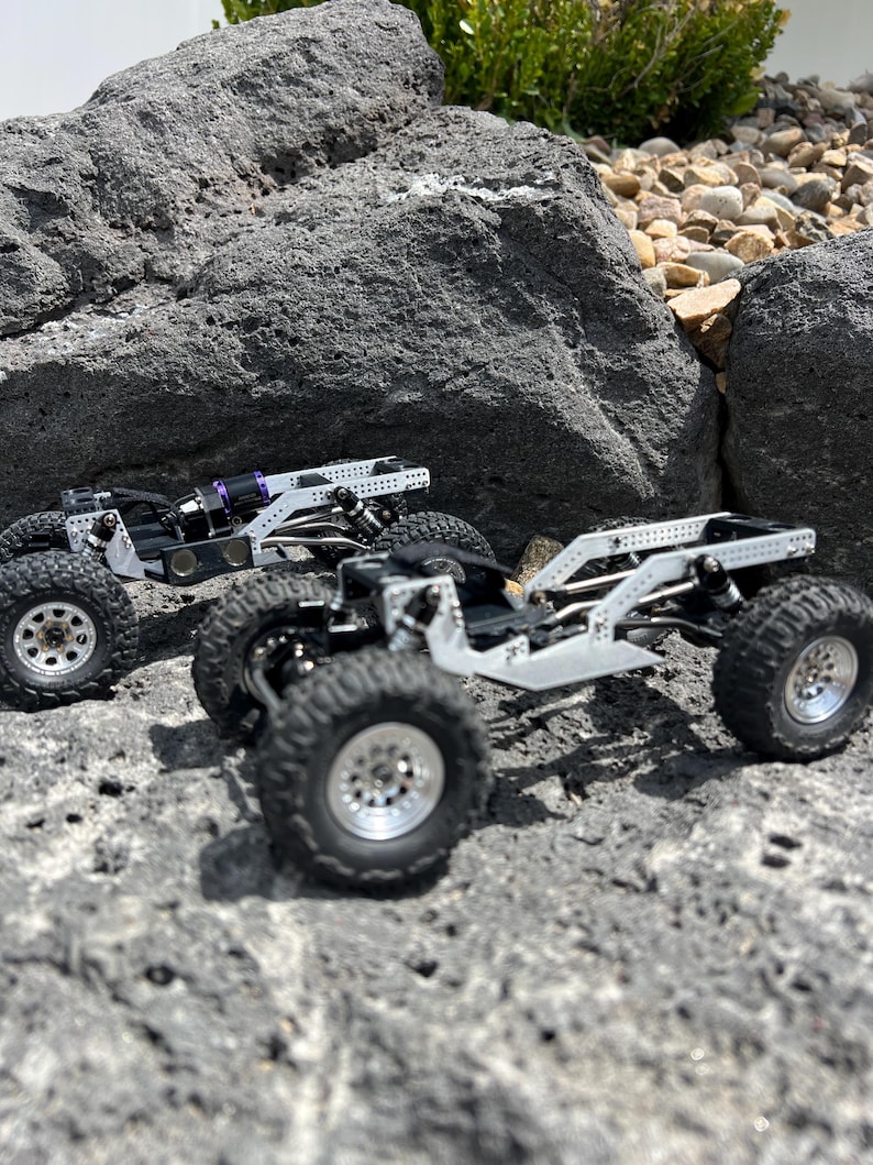 May include: Two miniature off-road vehicles with silver frames and black tires are positioned on a rocky surface. The vehicles have detailed suspension systems and silver wheels. One vehicle has a purple component. The scene is set outdoors with rocks and greenery.