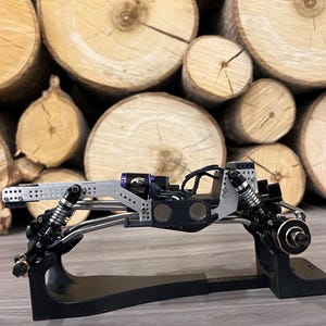 May include: A detailed, black and silver model car chassis with suspension and other mechanical components. The model sits on a black stand, with a backdrop of stacked, cut tree logs.