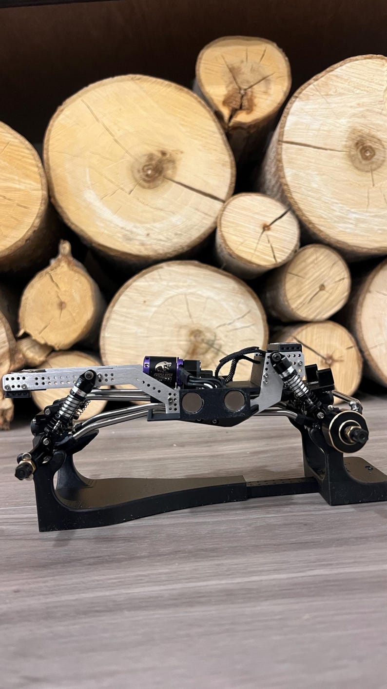 May include: A detailed, black and silver model car chassis with suspension and other mechanical components. The model sits on a black stand, with a backdrop of stacked, cut tree logs.